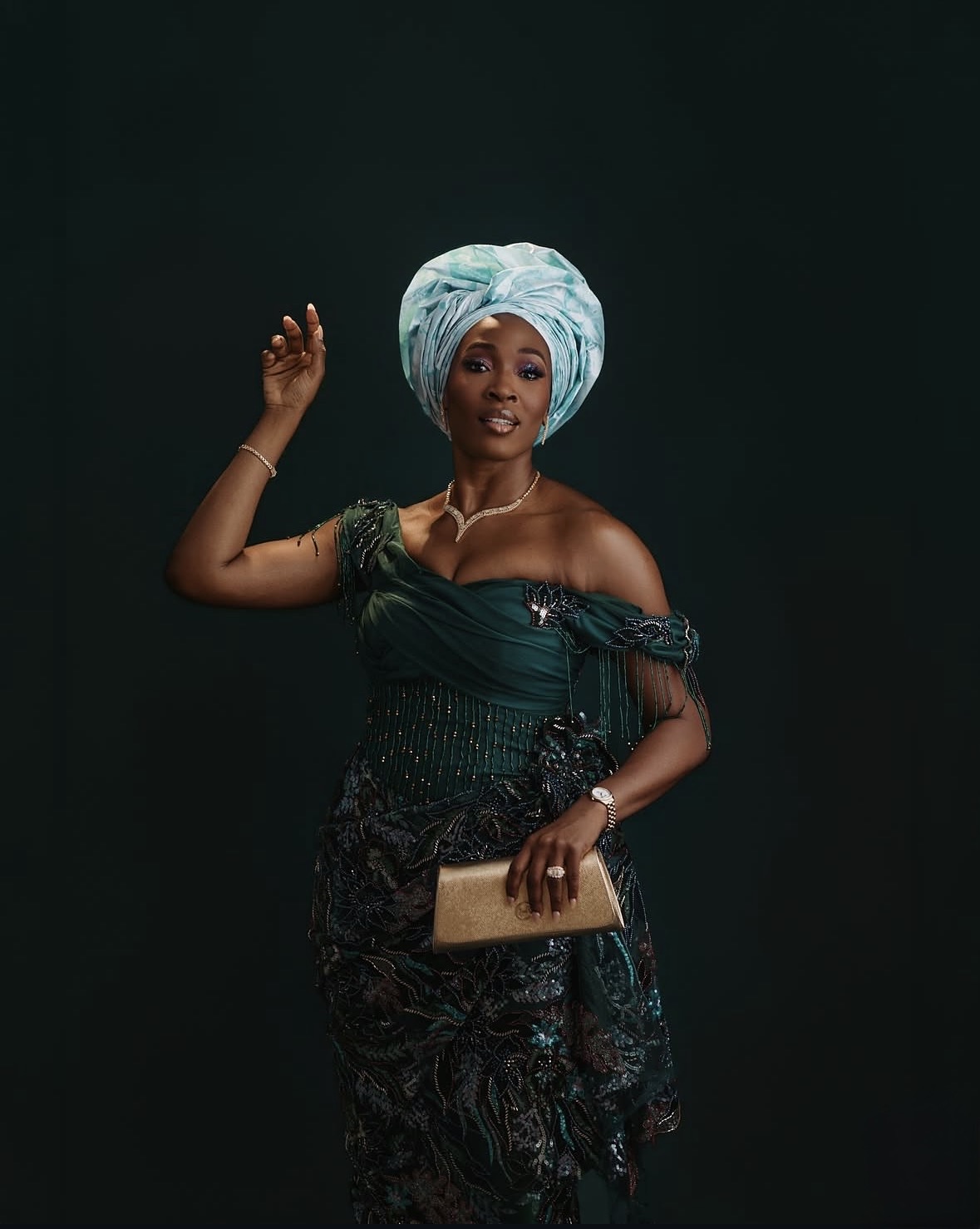 Tope Olowoniyan in a dark green embellished outfit and pale blue gele, holding a gold clutch
